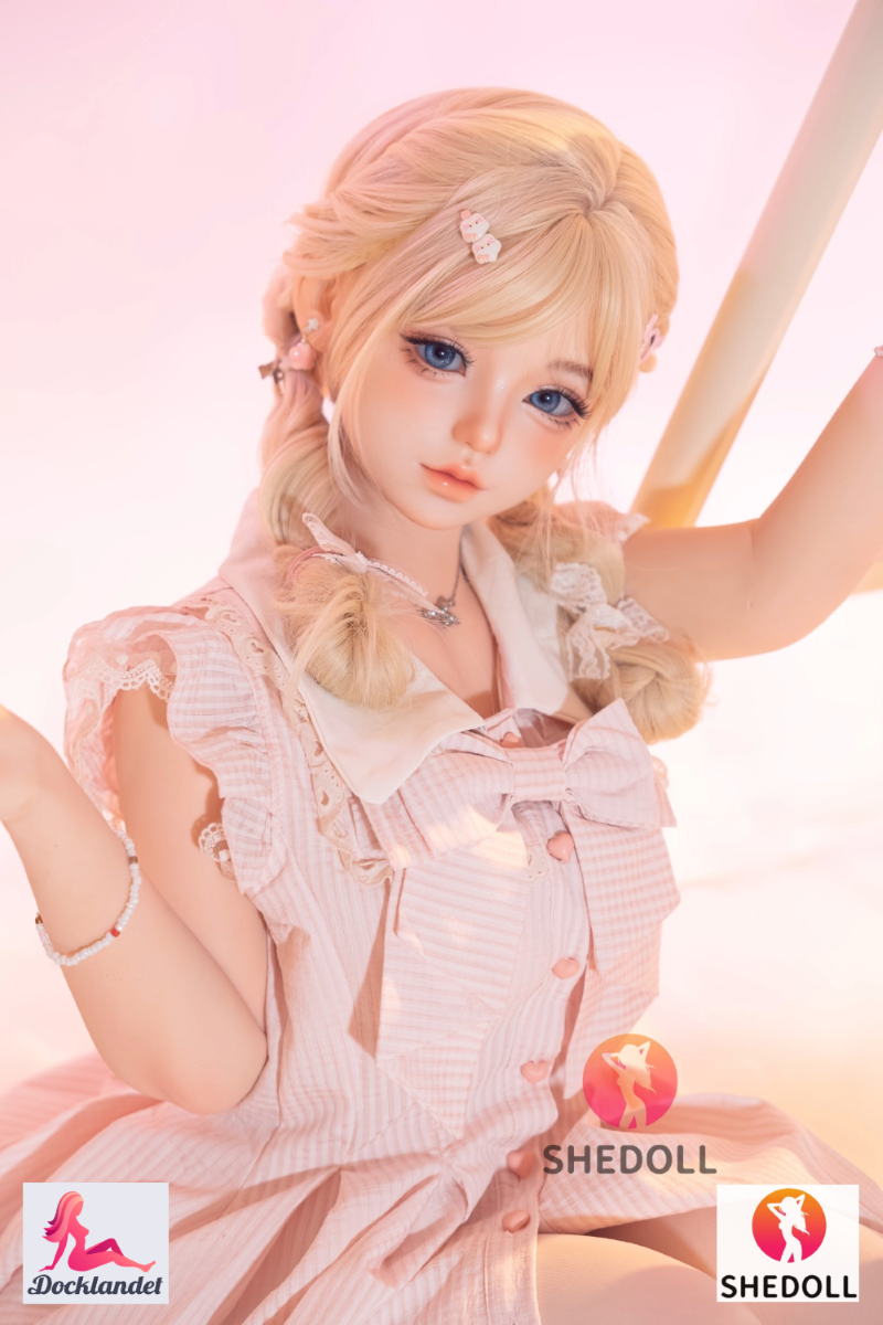 Sandy SHEDOLL 152 cm, sitter i rosa volangklänning, naturlig A-kupa, ljus pastellmiljö.